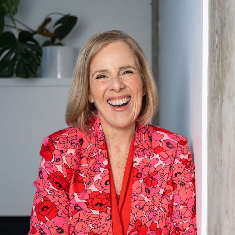 Michelle Ray. Headshot of Michelle Ray wearing a bright red floral top, posing inside with a potted plant behind her.