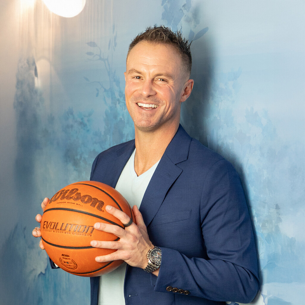 Alan Stein Jr. Headshot of Alan Stein Jr. wearing a navy suit jacket over a white t-shirt. He is posing with a basketball.
