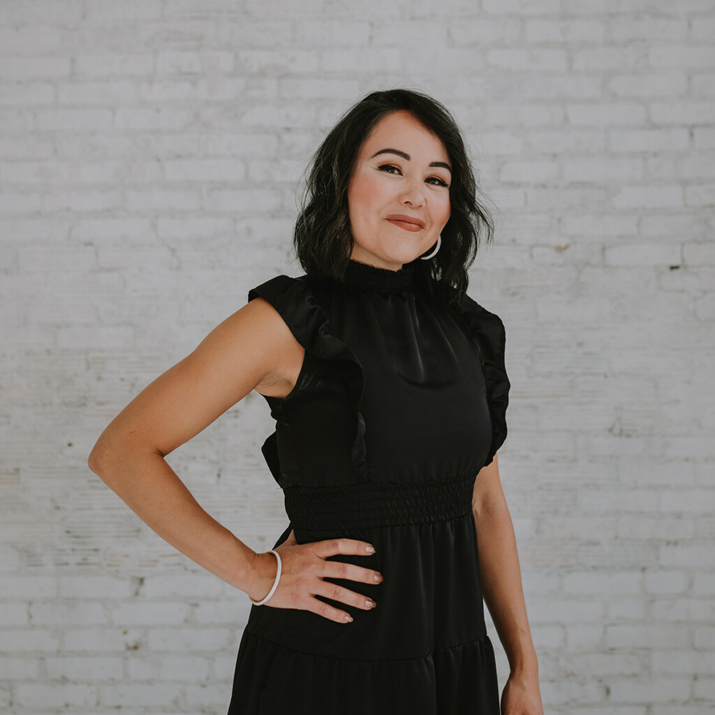 Christine Marie. Headshot of Christine Marie wearing a black short sleeved dress. She's posing with her hand on her hip in front of a white brick wall.