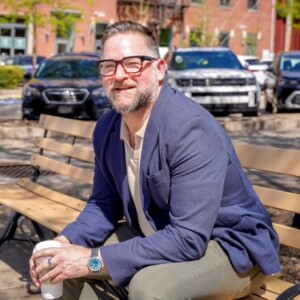 Headshot of keynote speaker, Jeff Siegler wearing a light-coloured shirt and blue suit jacket while sitting outside on a wooden park bench.