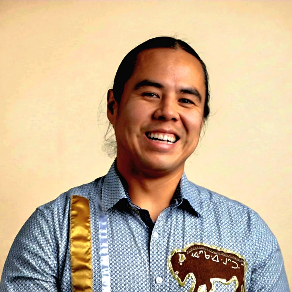 Headshot of John Langan in front of a yellow backdrop. He is smiling and is wearing a blue patterned shirt.