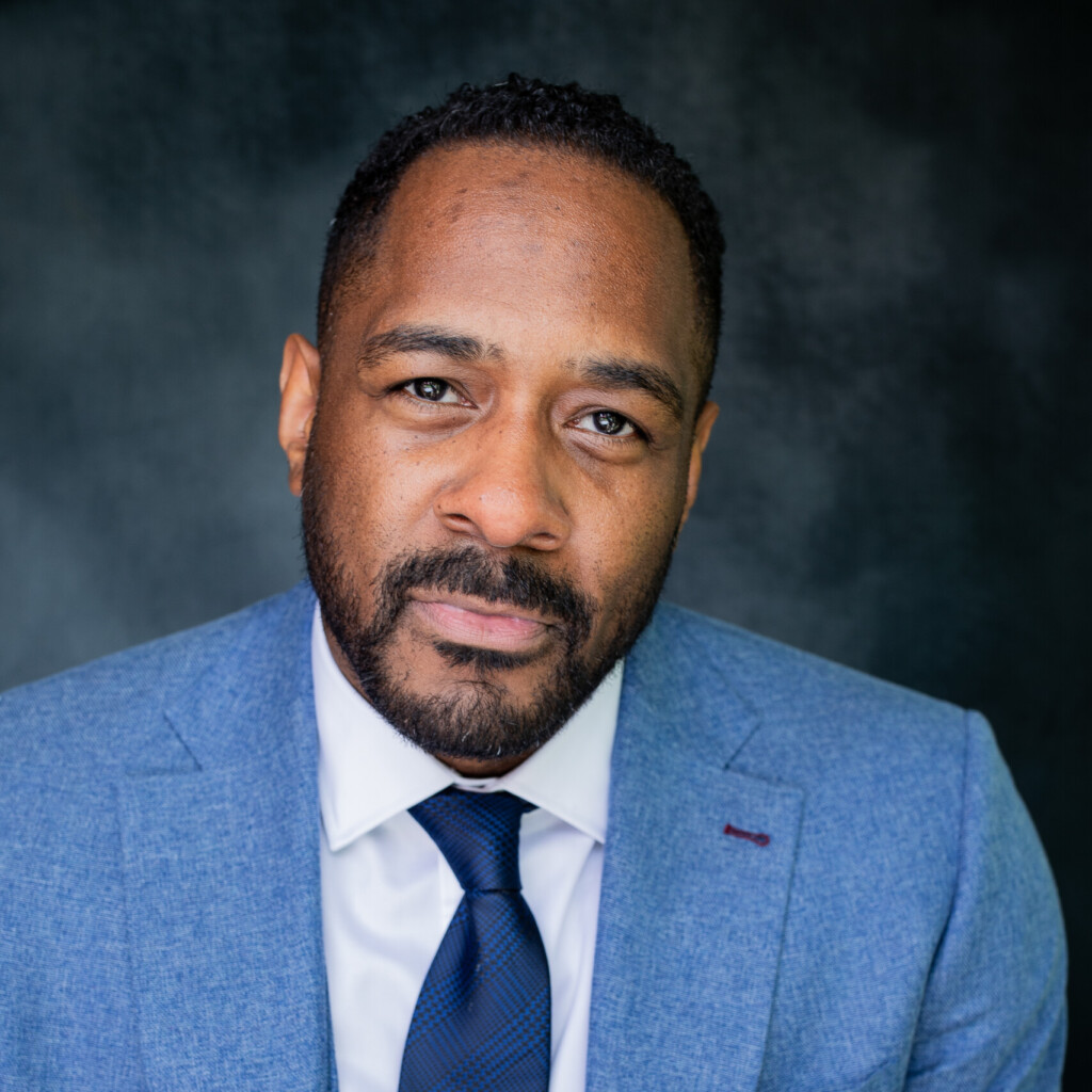 Professional headshot of Jesse Lipscombe in front of a grey background, wearing a white shirt and blue blazer and tie.