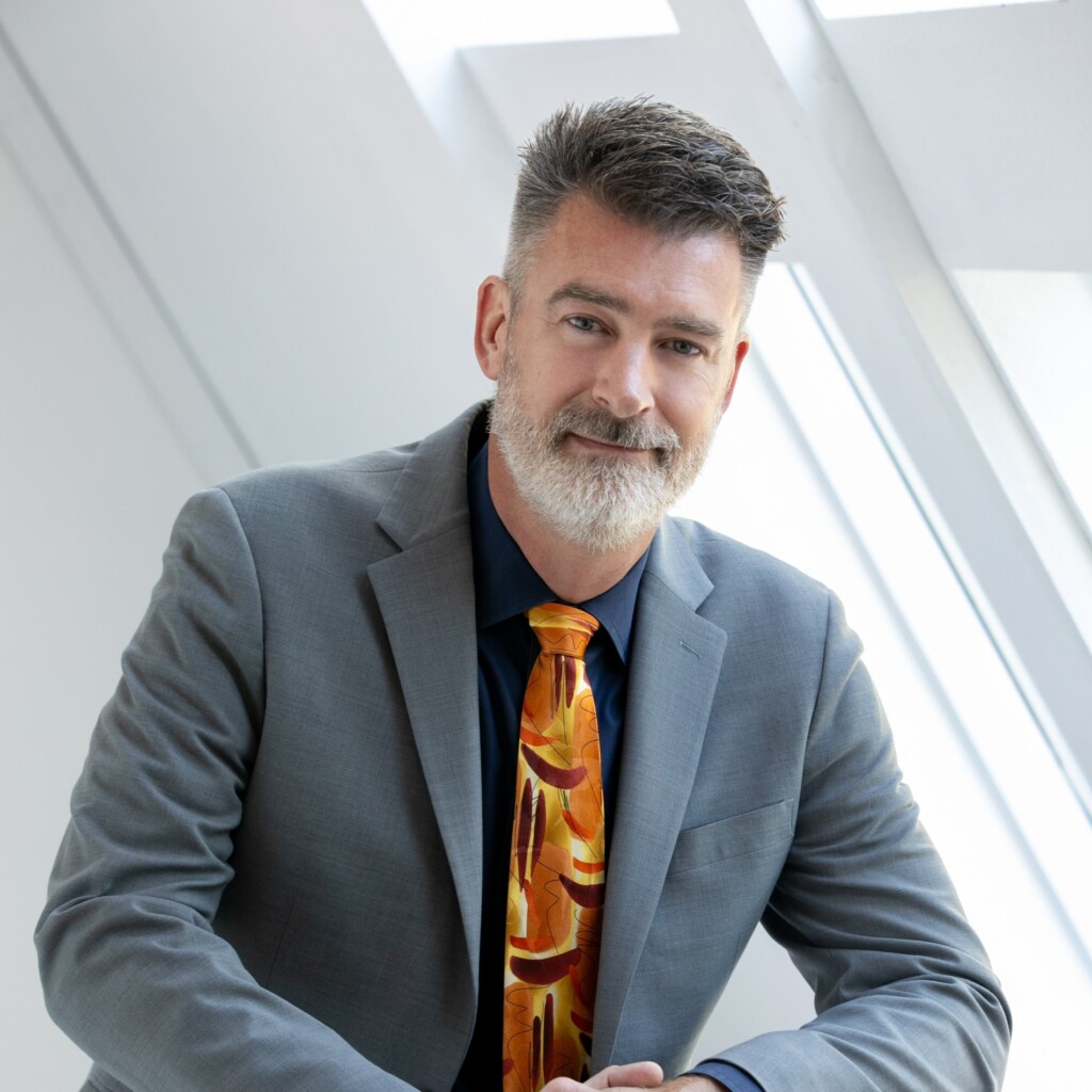Headshot of Peter Zeihan, wearing a grey blazer, and patterned orange neck tie, looking at the camera with his hands resting on the back of a black chair.