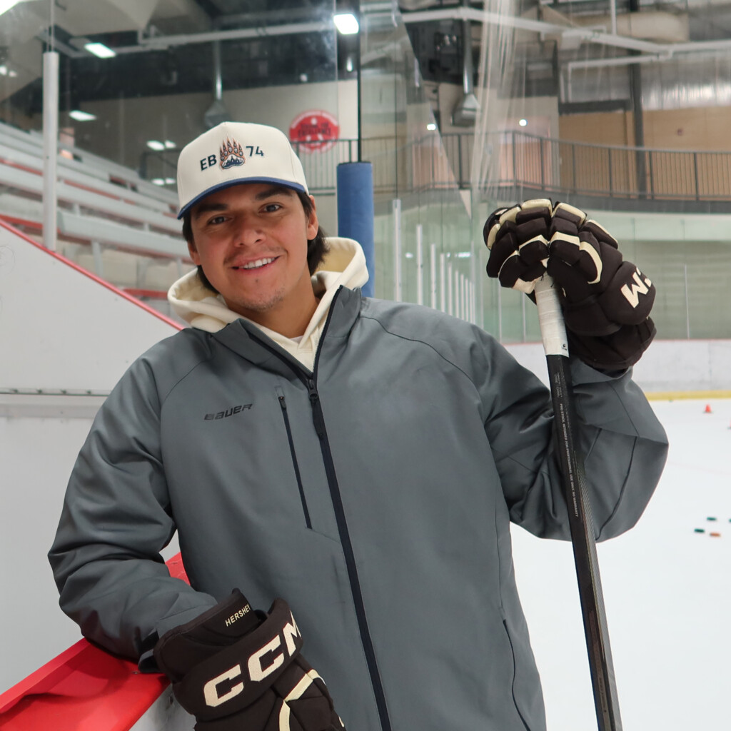 Ethan Bear. Headshot of Ethan bear posing in a hockey arena wearing a grey jacket, ball cap and hockey gloves.