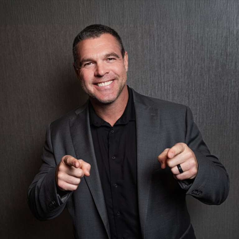 Jason Cyrus. Headshot of Jason Cyrus wearing a dark grey suit jacket over a black top. He's pointing at the camera and standing in front of a dark grey wall.
