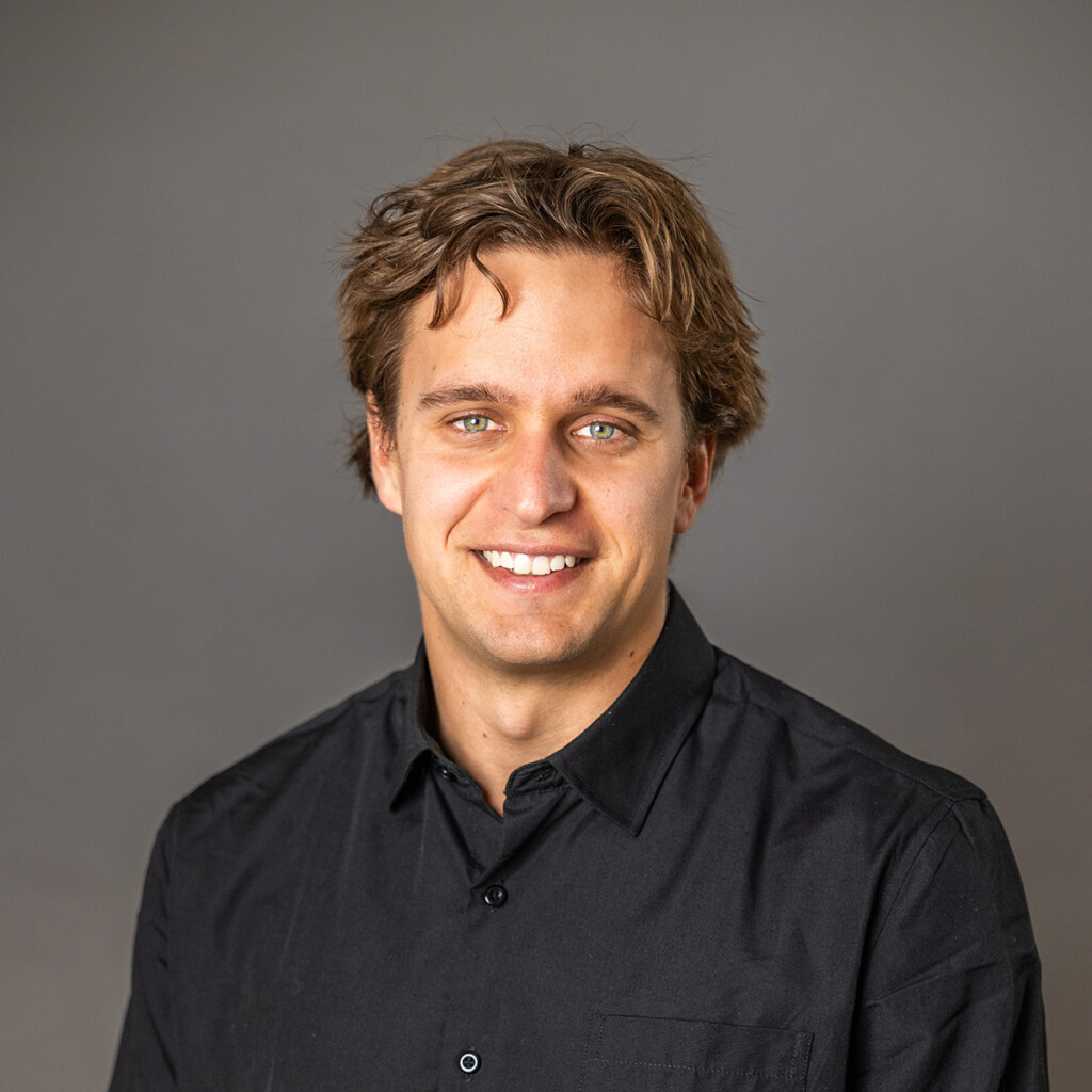 Lucas Arsenault Headshot. Lucas wearing a black collared shirt, sitting down in front of a grey studio backdrop.