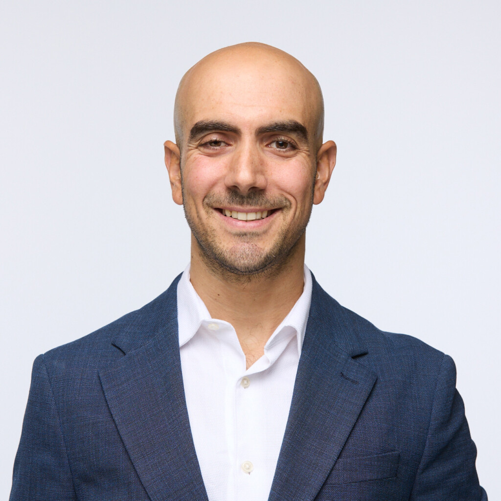 Headshot of Joze Piranian wearing a blue suit jacket over a white shirt. He's posing in front of a bright white back drop.