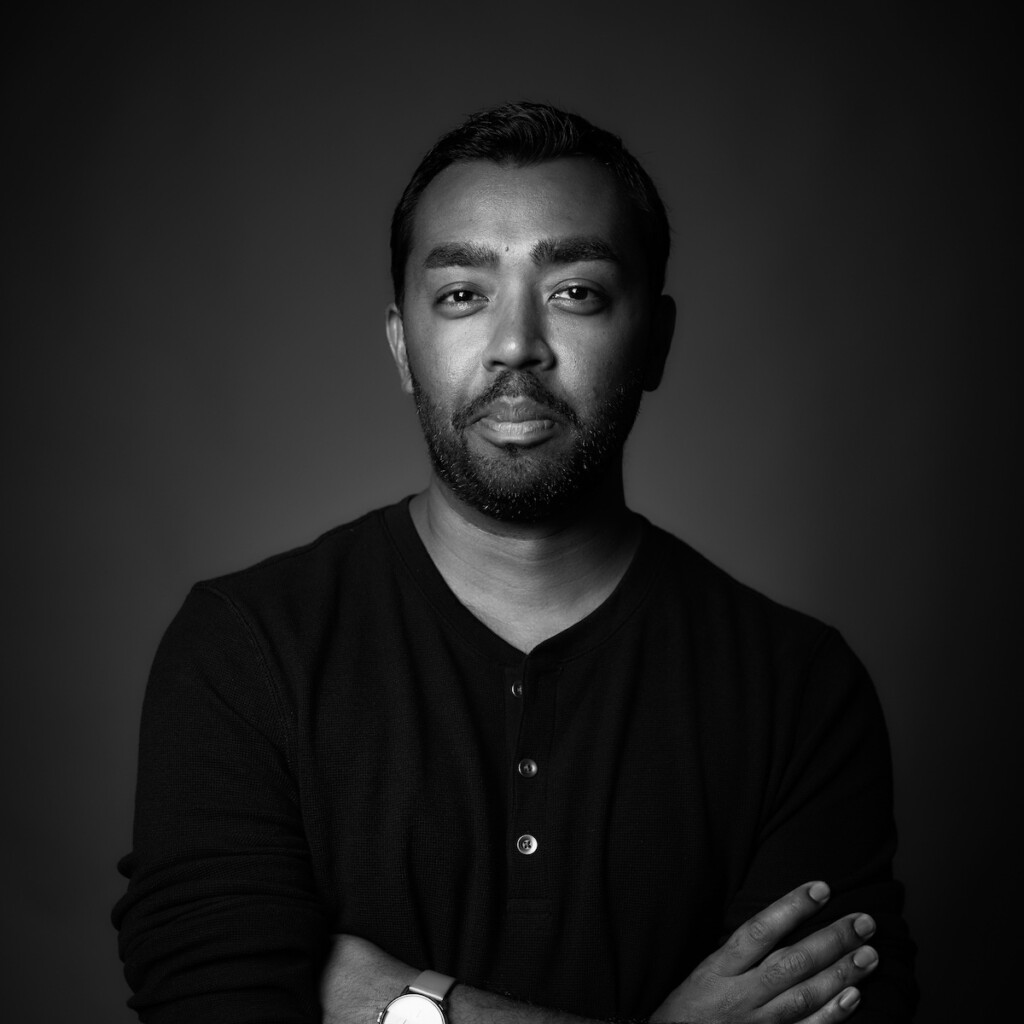 Suresh Doss headshot in black and white high-key studio lighting with a dark gradient backdrop. He's standing with his arms crossed in front of him.