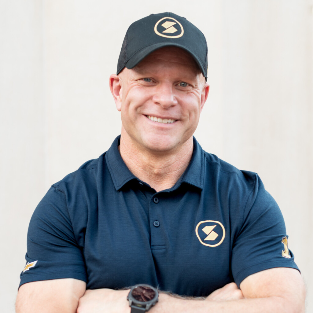 Professional headshot of Shaun Peet, keynote speaker, and Pit Crew Coach. He is smiling, standing with his arms crossed and is wearing a black polo shirt and baseball cap.