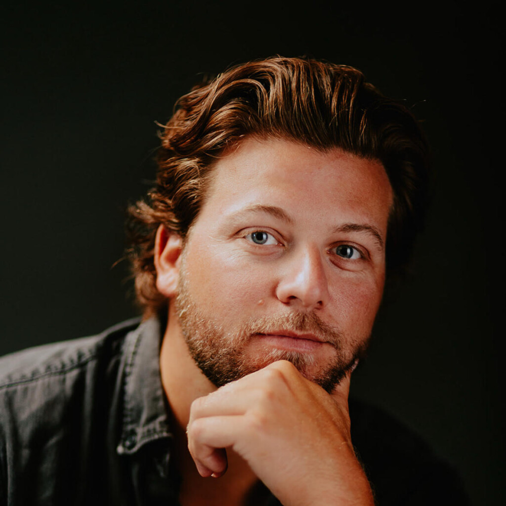 Jake Ernst headshot in front of a black background. He is wearing a black button up shirt and is resting his chin on his hand.