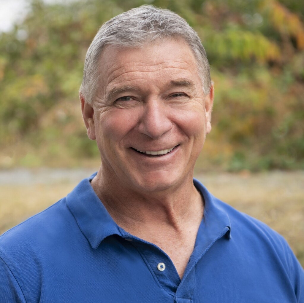 Rick Hansen. Headshot of Rick Hansen smiling, wearing a blue polo shirt, in front of a blurred, outdoor background.