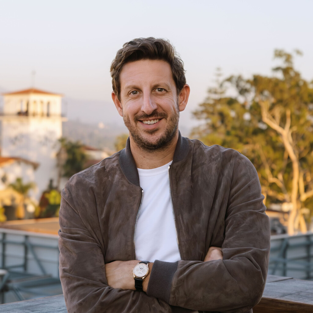 Zack Kass Headshot taken outside. He is posing with his arms crossed, wearing a brown jacket and white shirt.