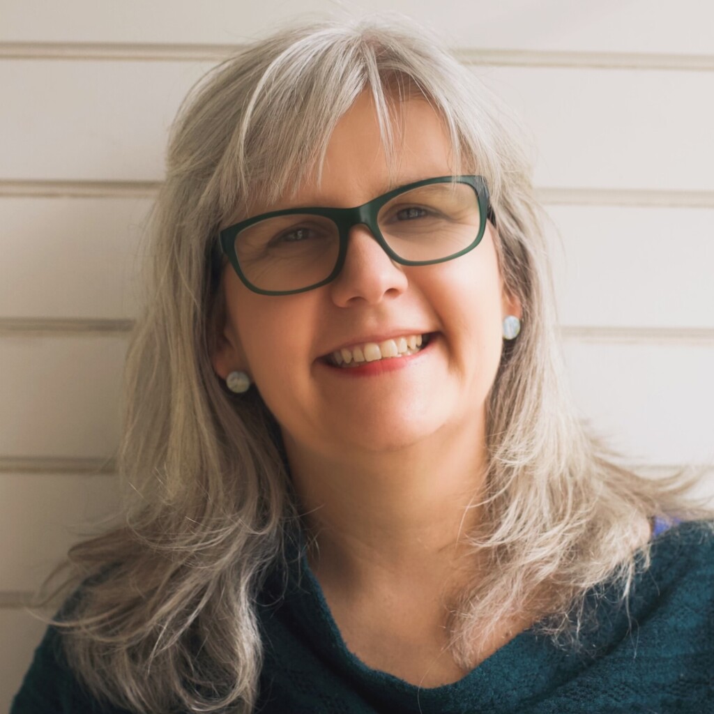 Headshot of Kate Robson, author and speaker. She is wearing glasses and a dark green knit sweater. She is standing in front of a white, panelled wall.