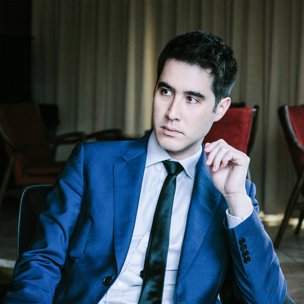 Headshot of David Kwong from the waist up. He is sitting in a chair, slightly leaning over as his elbow rests on the armrest of his chair. He is wearing a white collared shirt under a blue blazer with a black tie.