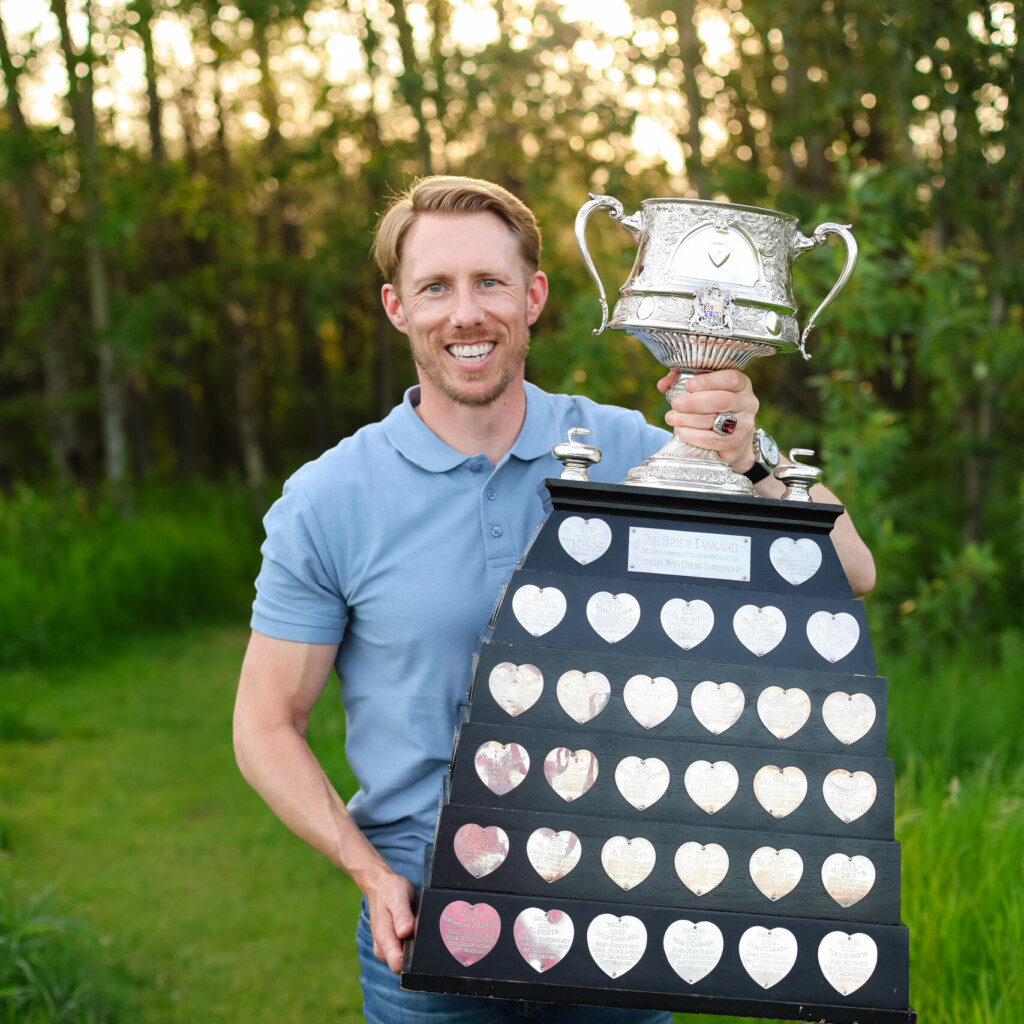 Headshot of Marc Kennedy, holding a large trophy, outdoors in front of a grassy, forest background.