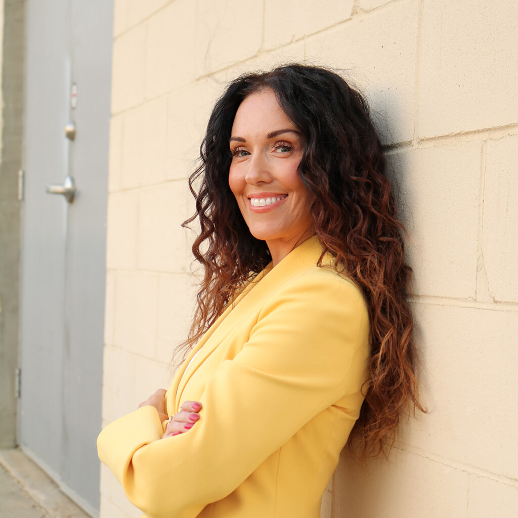 Jess Tetu. Professional headshot of Jess Tetu wearing a yellow jacket and posing outside in front of a beige wall.