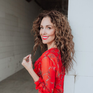 Jess Tetu. Professional headshot of Jess Tetu wearing a red dress and posing outside in front of a brick wall.