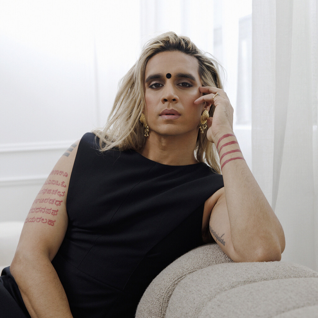 Vivek Shraya. Headshot of Vivek Shraya sitting on a white couch, wearing a black top in a brightly lit room.