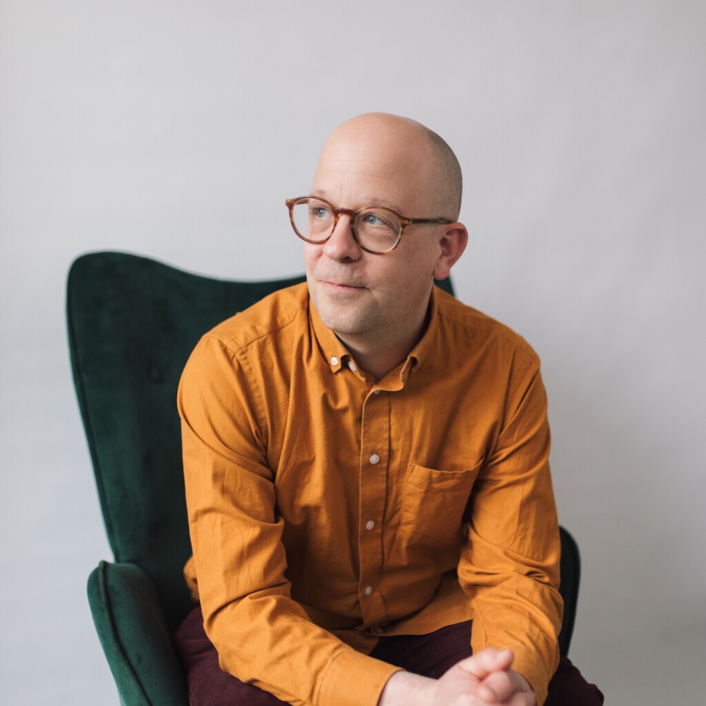 Headshot of Kevan Gilbert wearing a yellow button up shirt, and looking off into the distance as he is sitting in a green armchair.