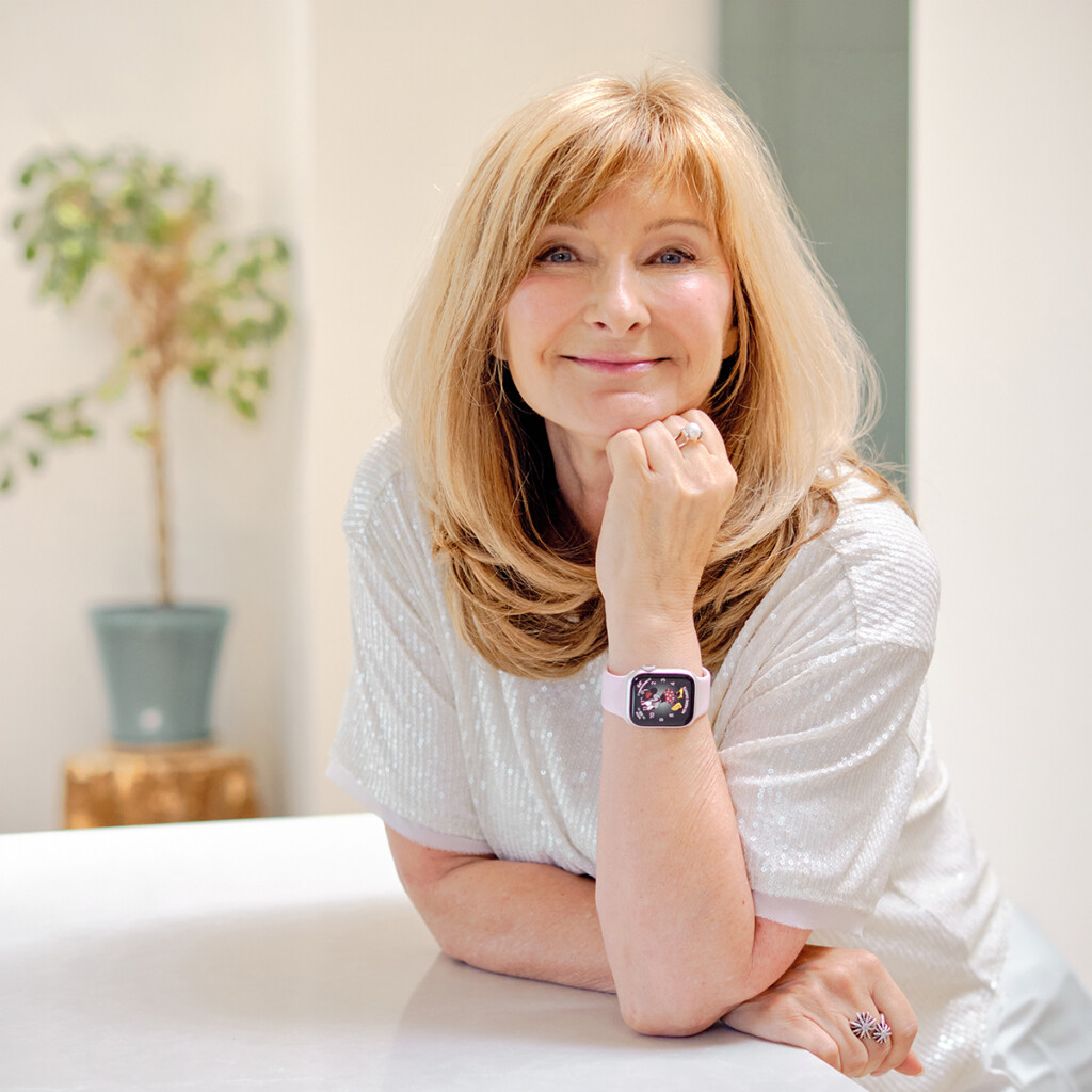 Headshot of Cheryl Cran wearing a shimmery white top, posing with her hand under her chin, leaning on a white counter.