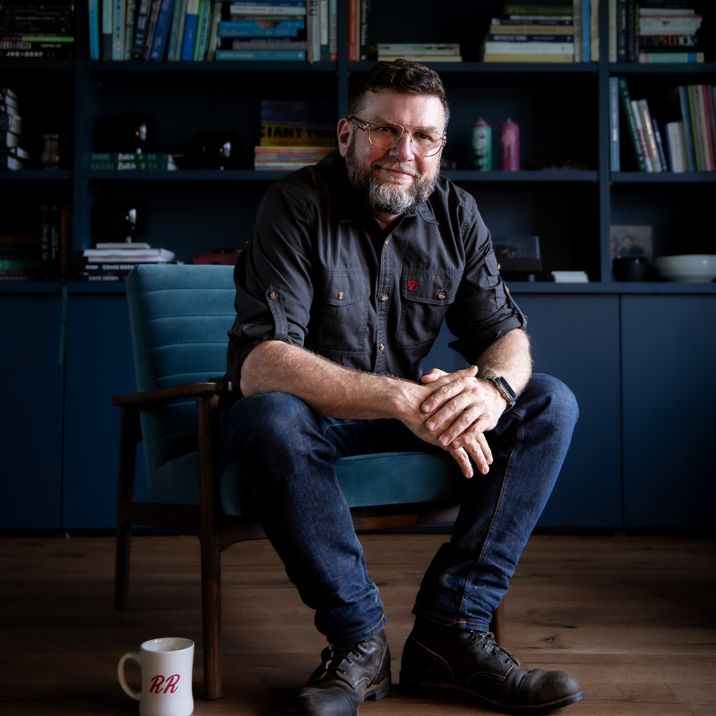 Headshot of Joel MacCharles wearing a dark collared shirt and jeans. He's sitting in a chair in front of a bookshelf.